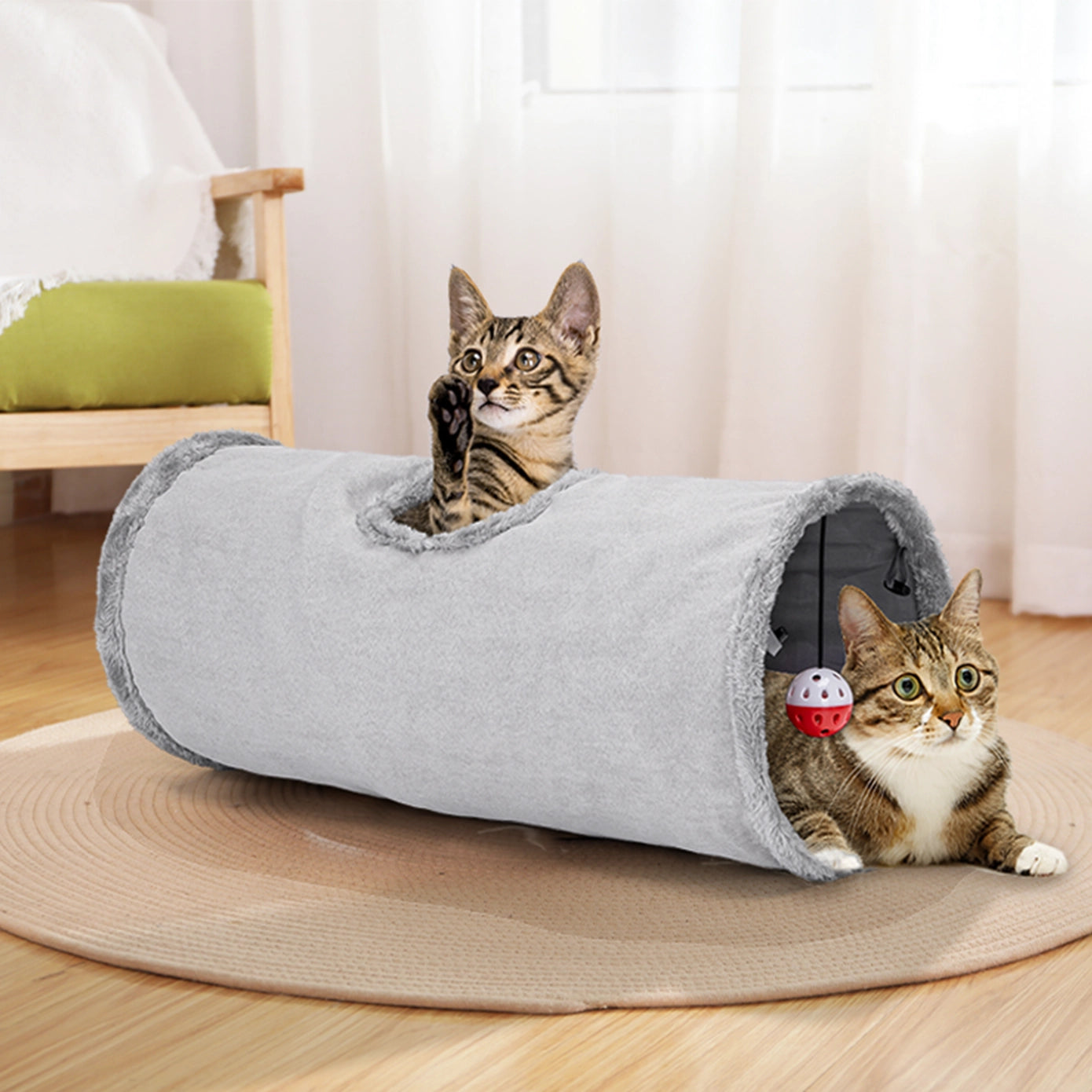 Tunnel pour chat en tissu gris doux, doté d'une boule rouge suspendue à l'intérieur. Deux chats jouent dedans, montrant son utilisation comme espace de jeu et de repos.